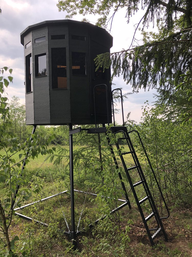 360 Pro hunting blind in black, elevated on a metal frame with a ladder, surrounded by greenery.