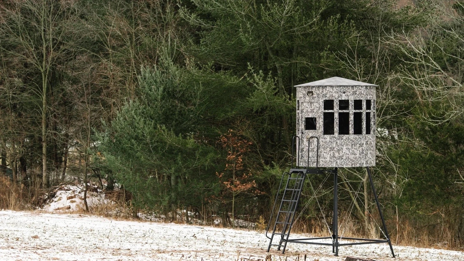 Elevated hunting blind with camouflage exterior in a snowy field surrounded by trees, side view.